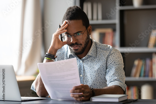 African American man wearing glasses dissatisfied by bad news received in letter, holding document, sitting at work desk, unexpected debt, bank or job dismissal notification, eviction notice