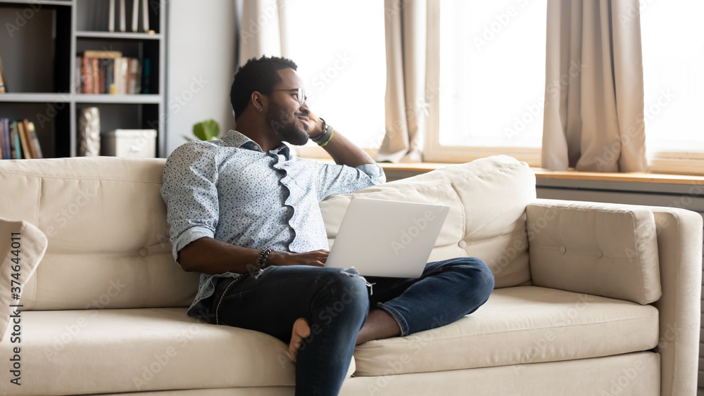 Smiling dreamy African American man sitting on couch with laptop, dreaming about good future, satisfied thoughtful businessman freelancer student pondering new opportunities or project strategy