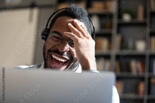 Laughing African American man wearing headphones looking at laptop screen close up, positive young male wearing glasses chatting online, making video call, watching funny movie or playing game