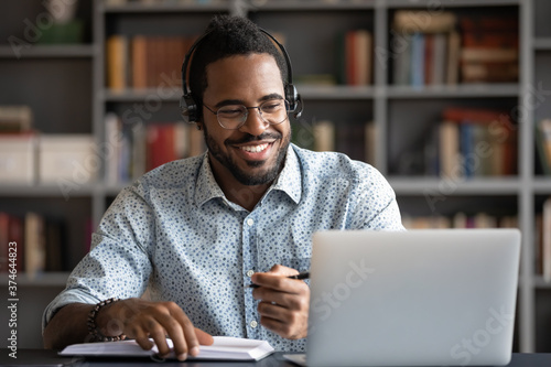 Smiling African American man wearing headphones looking at laptop screen, motivated student writing notes during online lesson, watching webinar, learning language online, sitting at work desk photo