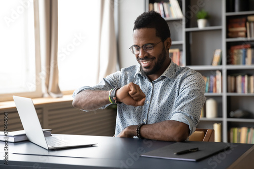Smiling African American businessman wearing glasses checking time, looking at wristwatch on hand, positive happy young man entrepreneur sitting at desk, planning work, task management concept