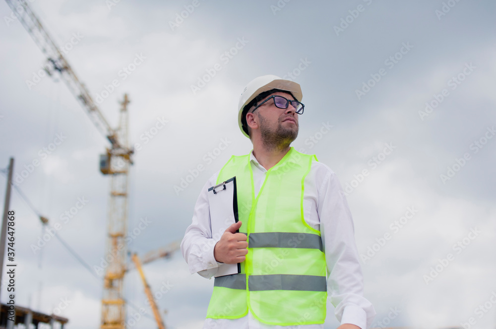 Chief in a vest and a construction helmet with documents in his hands, against the background of a tower crane and the sky with clouds. House construction inspection. Chief in a vest and a construction helmet with documents in his hands, against the background of a tower crane and the sky with clouds. House construction inspection.