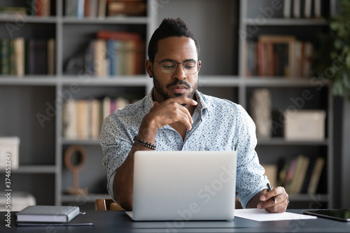 Thoughtful African American businessman looking at laptop screen, touching chin, pondering project plan or strategy, creative ideas, freelancer working online, sitting at desk in modern cabinet