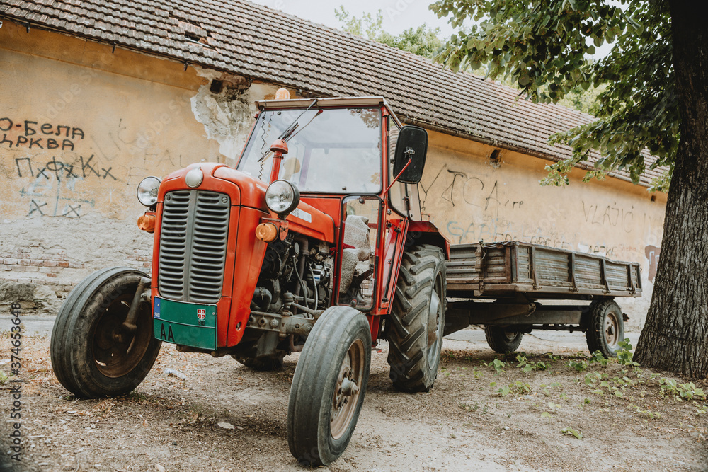 Obraz premium One old red tractor with trailer standing alone in front of building on the gravel street in shadow under the tree during the summer sunny day in Serbia
