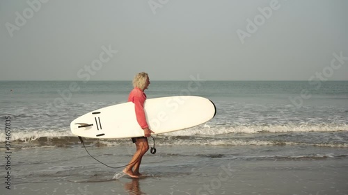 An old man walks along the sea beach with a surfboard. An active pensioner goes in for sports. Grandpa is a surfer