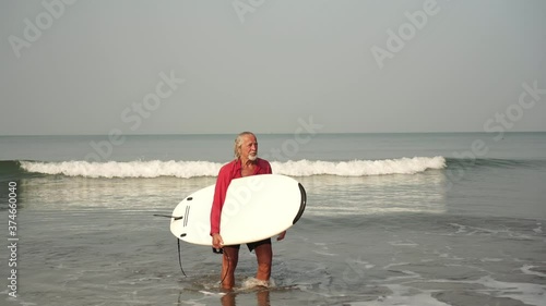 An old man comes out of the sea with a surfboard. A senior pensioner goes in for sports. The elderly person surfs and leads an active sports lifestyle