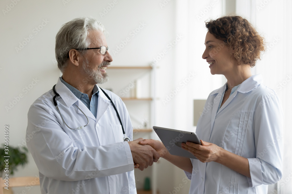 Smiling mature doctor wearing glasses shaking young nurse hand, thanking for help, good work result, friendly woman therapist holding digital tablet, greeting colleague, teamwork concept Smiling mature doctor wearing glasses shaking young nurse hand, thanking for help, good work result, friendly woman therapist holding digital tablet, greeting colleague, teamwork concept
