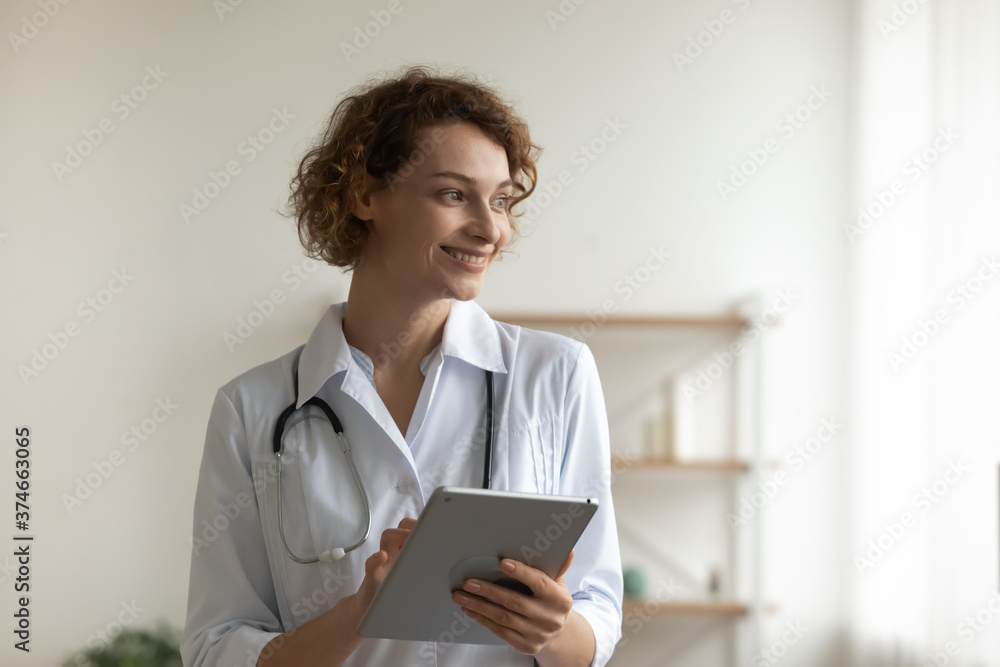 Close up smiling young woman wearing uniform with stethoscope, doctor holding digital tablet, pondering ideas, looking to aside, standing in hospital office, dreaming about opportunities, future Close up smiling young woman wearing uniform with stethoscope, doctor holding digital tablet, pondering ideas, looking to aside, standing in hospital office, dreaming about opportunities, future