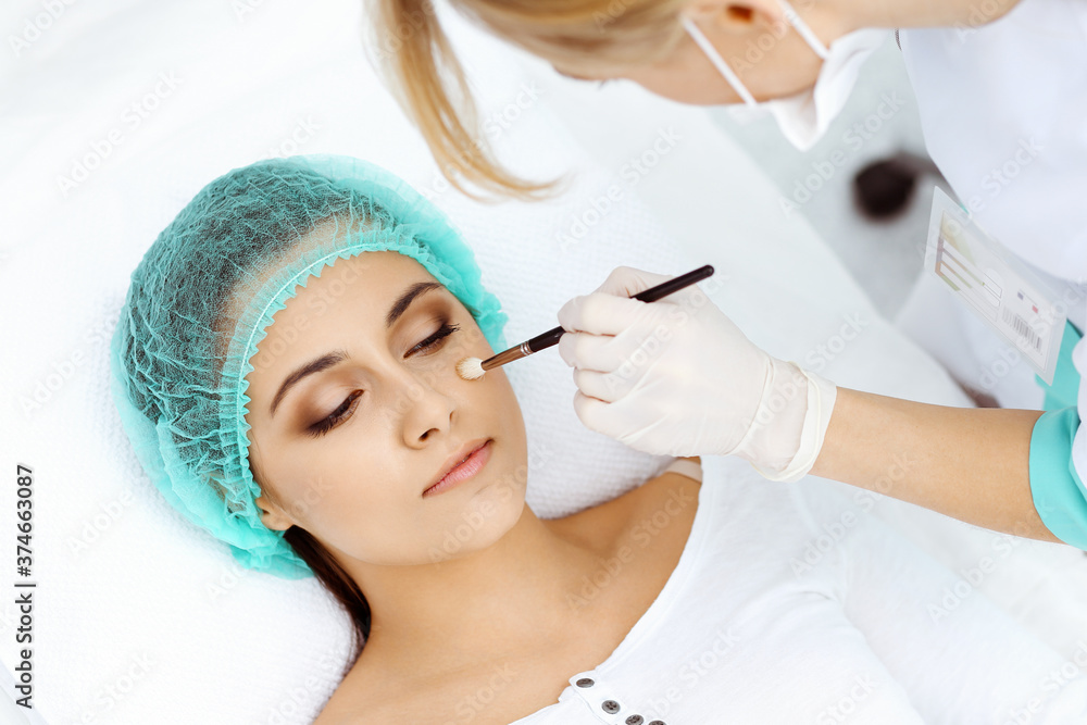 Professional beautician is doing cosmetic procedure at light medical background touching patient's face with brush, closeup. Cosmetology treatment Professional beautician is doing cosmetic procedure at light medical background touching patient's face with brush, closeup. Cosmetology treatment