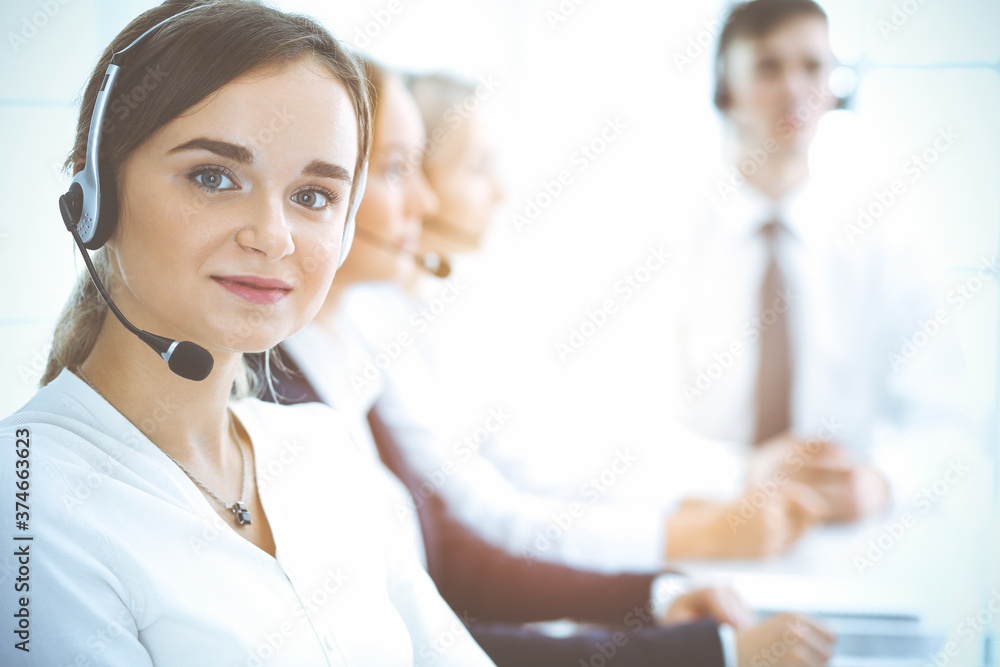 Cheerful smiling business woman with headphones consulting clients. Group of diverse phone operators at work in sunny office.Call center and business people concept Cheerful smiling business woman with headphones consulting clients. Group of diverse phone operators at work in sunny office.Call center and business people concept