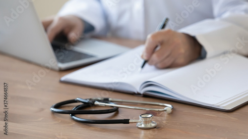 Close up stethoscope, mature doctor filling documents and using laptop, sitting at work desk in hospital, senior therapist practitioner gp writing illness history, prescription in patient card