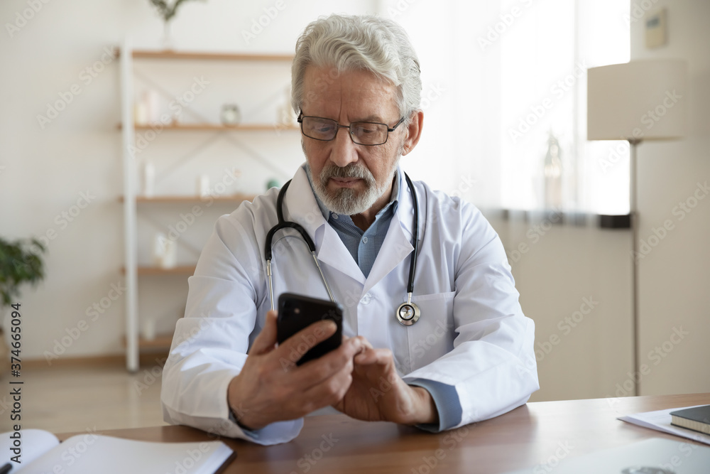 Serious mature doctor wearing glasses and white coat uniform with stethoscope looking at smartphone screen, sitting at work desk in hospital, reading email, chatting with patient, healthcare Serious mature doctor wearing glasses and white coat uniform with stethoscope looking at smartphone screen, sitting at work desk in hospital, reading email, chatting with patient, healthcare