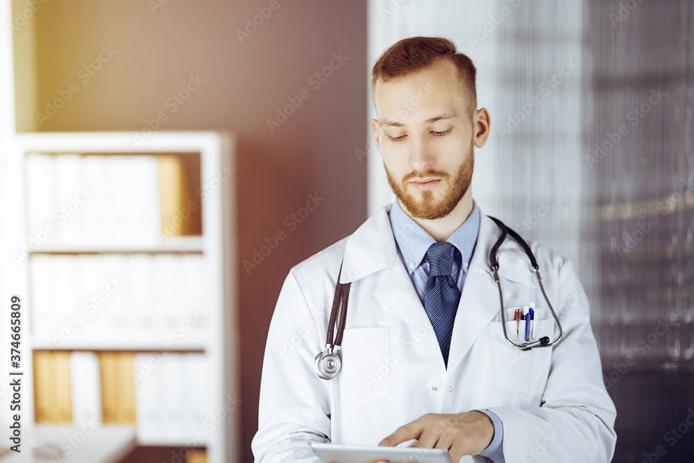 Friendly red-bearded doctor using tablet computer in sunny clinic at his working place. Medicine concept Friendly red-bearded doctor using tablet computer in sunny clinic at his working place. Medicine concept