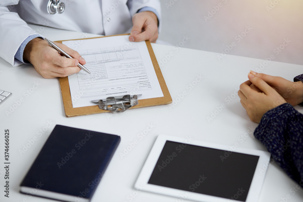Fototapeta premium Friendly red-bearded doctor and patient woman discussing current health examination while sitting in sunny clinic. Medicine concept