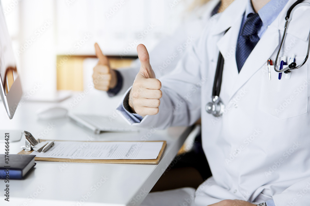 Two doctors sitting with thumbs up in hospital office. Medical help, countering viral infection and medicine concept Two doctors sitting with thumbs up in hospital office. Medical help, countering viral infection and medicine concept