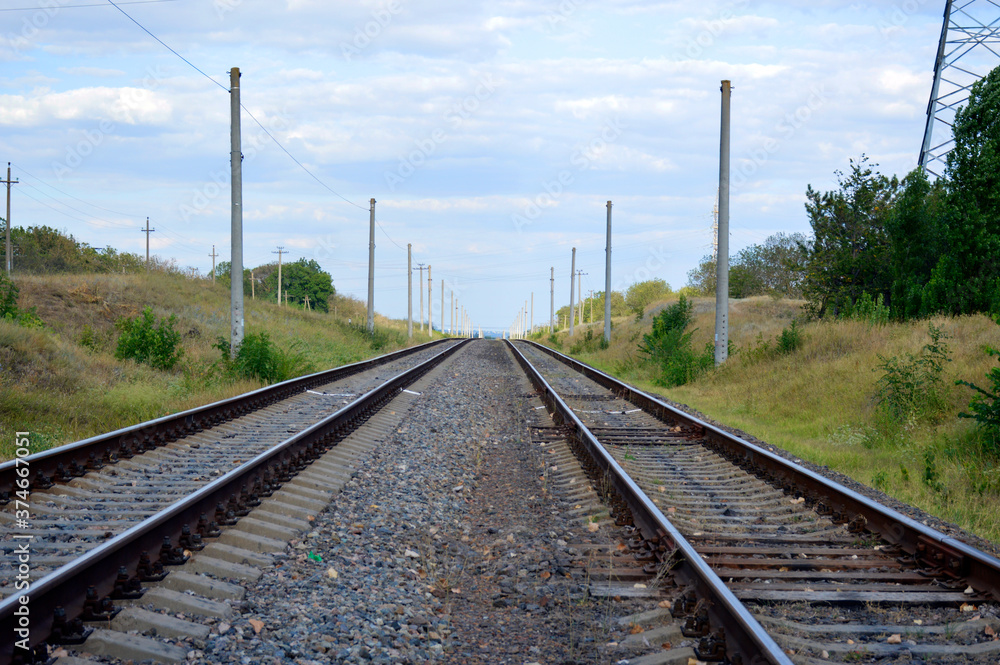 two railway tracks go into the distance, trees grow along the edges, and above there is a blue sky with white clouds