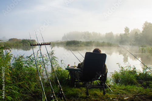 A man fisherman sits on the shore of a forest lake in a chair at dawn
