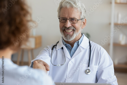 Close up smiling mature doctor wearing glasses and white uniform with stethoscope shaking female patient hand, greeting at medical appointment, making health insurance agreement, gratitude