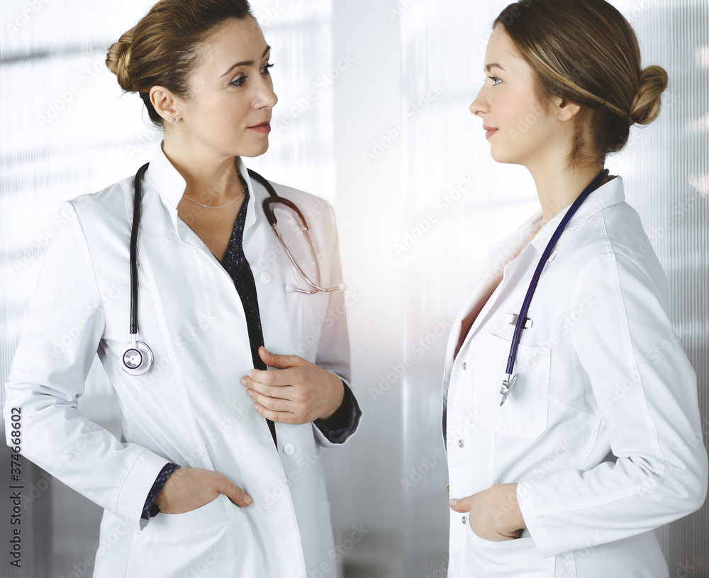 Two female physicians are discussing medical therapy, while standing at the table in a sunny clinic office. Doctors use pc computer at work. Teamwork in medicine Two female physicians are discussing medical therapy, while standing at the table in a sunny clinic office. Doctors use pc computer at work. Teamwork in medicine