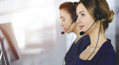 Two busineswomen have conversations with the clients by headsets, while sitting at the desk in a sunny modern office. Diverse people group in a call center