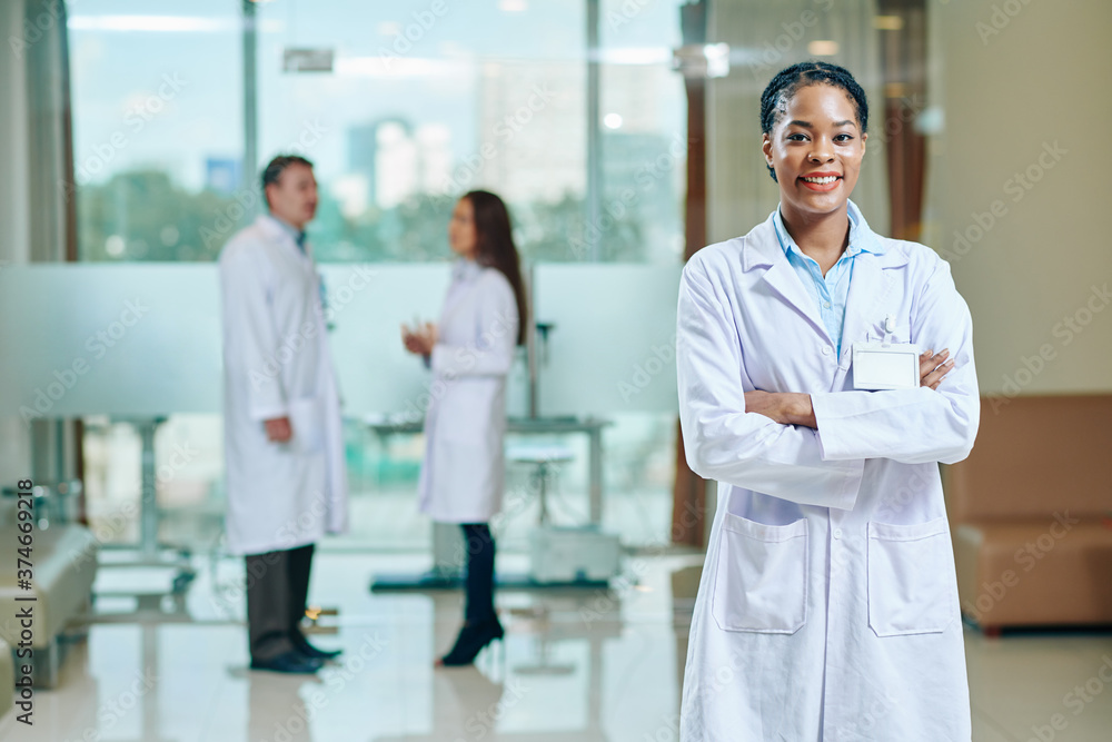 Portrait of pretty smiling young Black doctor in white labcoat standing in hall of clinic Portrait of pretty smiling young Black doctor in white labcoat standing in hall of clinic