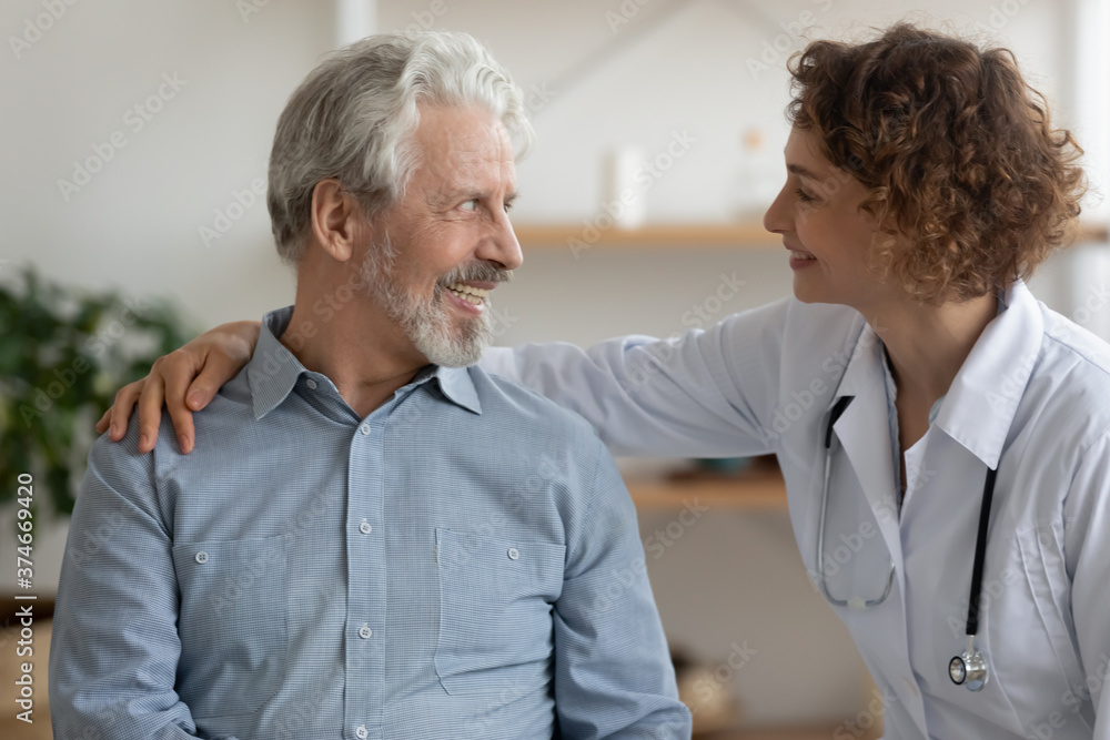 Obraz premium Close up friendly young woman nurse caregiver hugging smiling mature patient, caring doctor wearing white coat and stethoscope supporting happy senior man at meeting, healthcare concept