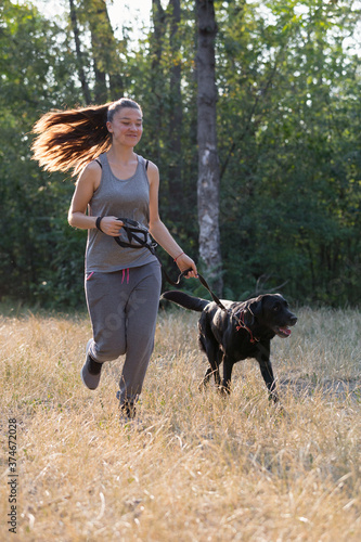 Young attractive sport girl running with black Labrador in park. Healthy lifestyle. Walk with favorite pet in wood.