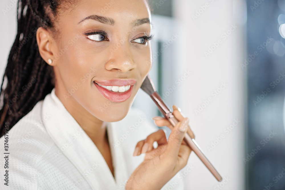 Beautiful young Black woman applying blush or face powder in front of mirror Beautiful young Black woman applying blush or face powder in front of mirror
