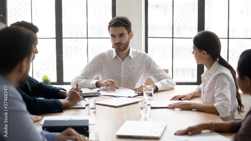 Confident young skilled businessman sitting at table, holding negotiations meeting with multiracial colleagues in office. Millennial male mentor instructing mixed race teammates at workplace.