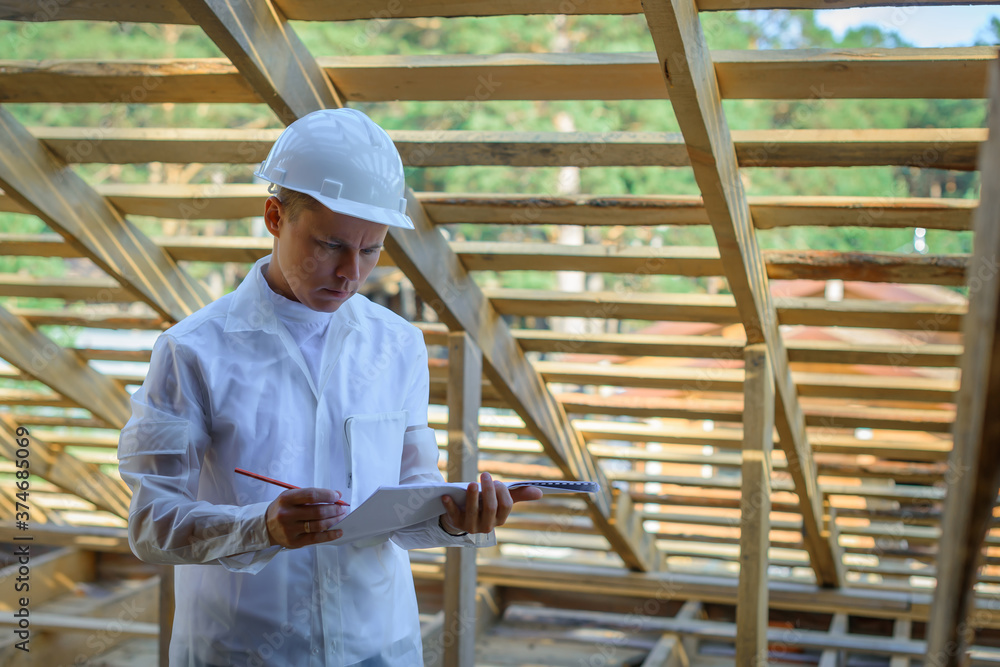 Foreman holding drawings, looking the project of wooden house, working on a construction site. Young attractive engineer in a white helmet checks the roof frame. Concept career architect. Copy space.