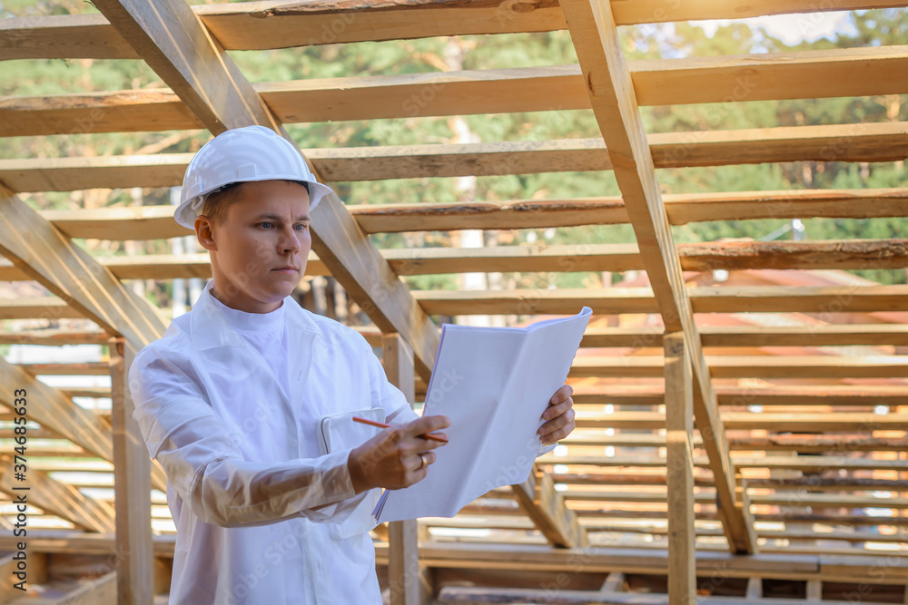 Foreman holding drawings, looking the project of wooden house, working on a construction site. Young attractive engineer in a white helmet checks the roof frame. Concept career architect. Copy space. Foreman holding drawings, looking the project of wooden house, working on a construction site. Young attractive engineer in a white helmet checks the roof frame. Concept career architect. Copy space.