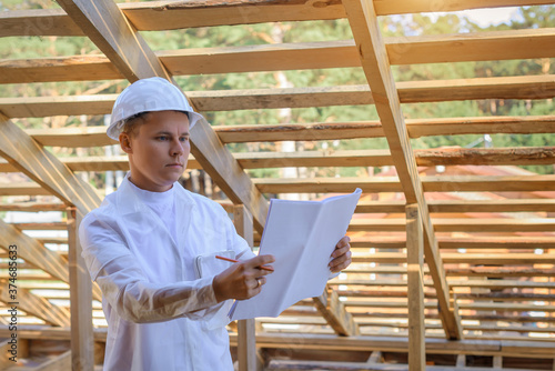 Foreman holding drawings, looking the project of wooden house, working on a construction site. Young attractive engineer in a white helmet checks the roof frame. Concept career architect. Copy space.