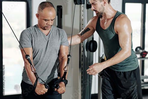 FItness trainer controlling posture of client doing exercise in cable crossover machine to strengthen muscles of arms and upper chest