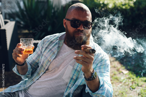 Portrait of an attractive bearded business man smoking a cigar with glass of whiskey in a summer cafe. photo