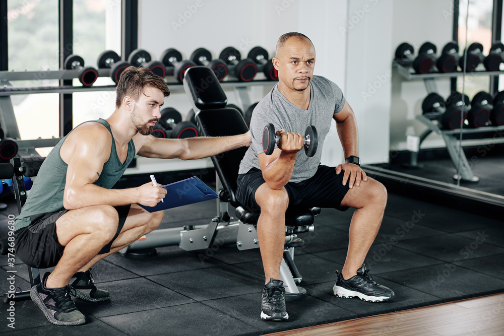 Personal trainer asking mature client to keep back straight when he is doing biceps exercise with dumbbell in gym