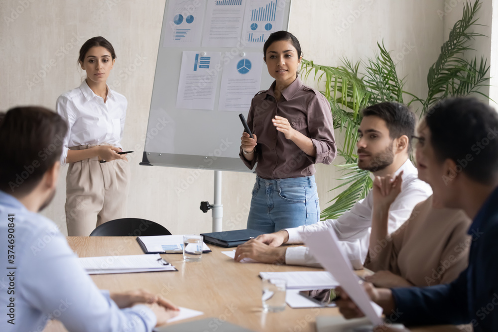 Fototapeta premium Focused young multiracial female speakers standing near flipchart with marketing research results, answering questions of educational conference participants, giving workshop seminar in office.