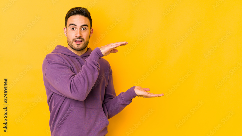 Young caucasian man isolated on yellow bakground shocked and amazed holding a copy space between hands.