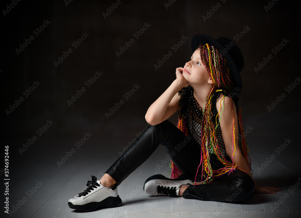 Teenager with african pigtails, in black hat, leggings, mesh blouse and sneakers. Sitting on floor, posing on dark background Teenager with african pigtails, in black hat, leggings, mesh blouse and sneakers. Sitting on floor, posing on dark background