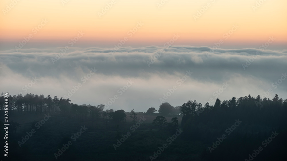 Fototapeta premium Majestic landscape image of cloud inversion at sunset over Dartmoor National Park in Engand with cloud rolling through forest on horizon