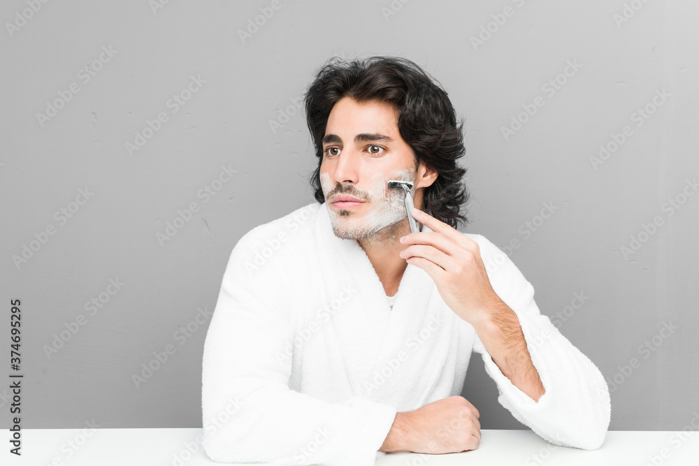 Young caucasian man shaving his beard isolated on a grey background