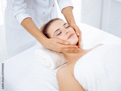 Young and blonde woman enjoying facial massage in spa salon. Beauty concept