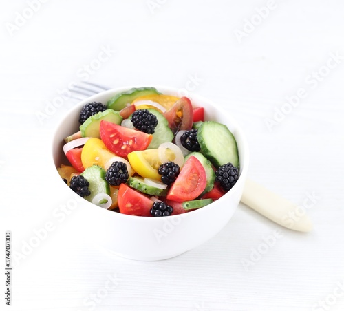 Vegetable berry salad of fresh yellow  red  black tomatoes  cucumbers  onions and blackberries in a bowl on a white background