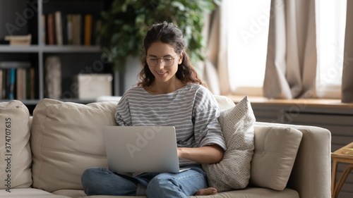 Pretty millennial smiling woman in glasses relaxing on couch with computer on laps, enjoying watching movie online, using dating application, communicating with friends in social networks, web surfing