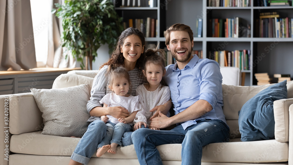 Portrait of full relaxed family with adorable little children, sitting on sofa. Happy young couple parents embracing small kids daughters, enjoying free leisure weekend time together in living room.
