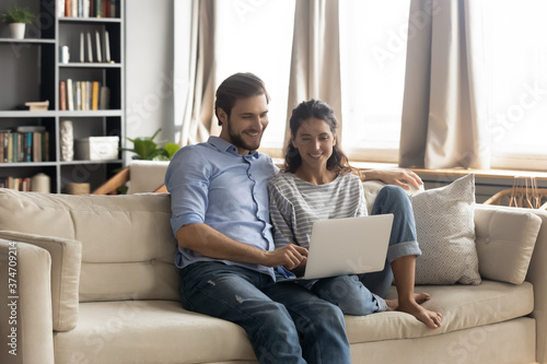 Happy young romantic bonding couple relaxing on comfortable sofa, watching online movie on computer, planning abroad vacation, booking tickets, shopping or web surfing information together at home.