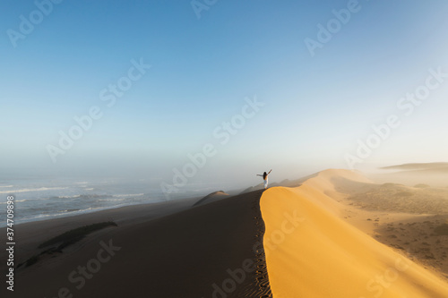 Woman walking on top of huge sand dune in Morocco Sahara desert. Beautiful warm sun light and mist in morning. View from behind. Freedom concept.