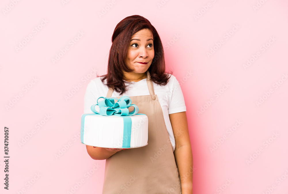 Middle age latin woman holding a cake isolated on a pink background confused, feels doubtful and unsure. Middle age latin woman holding a cake isolated on a pink background confused, feels doubtful and unsure.