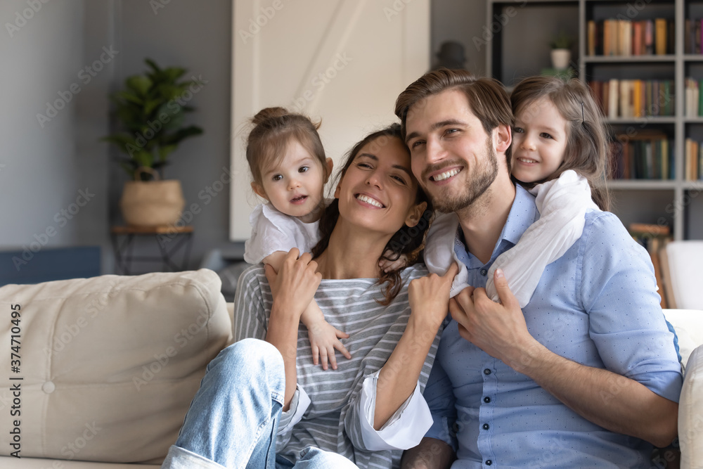 Loving small baby girls embracing smiling parents, looking away. Bonding affectionate young married couple enjoying spending holiday weekend free leisure time with two cute daughters, watching tv.