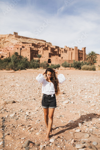 Young curly woman eastern appearance walking on background of kasbah Ait-Ben-Haddou. Travel in Morocco, Ouarzazate. Summer vacations, travel lifestyle concept. © Oleg Breslavtsev