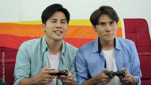 Young man with boyfriend holding wireless controller for play the games, they are sitting near red couch with the flag of LGBT Gender diversity symbol. They are having fun smile and happy together. 
